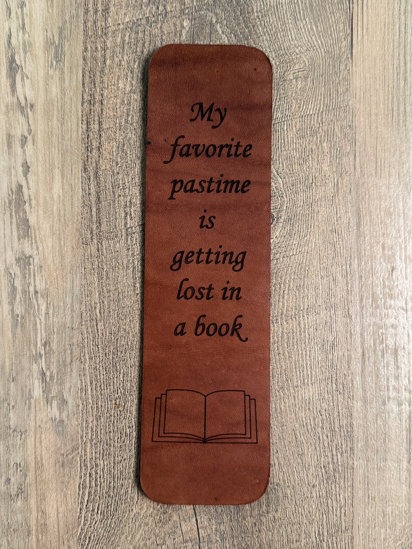 A brown, rectangular leather bookmark with rounded corners. Laser-engraved with the quote "My favorite pastime is getting lost in a book. " A laser-engraved outline of an open book is below the quote text.