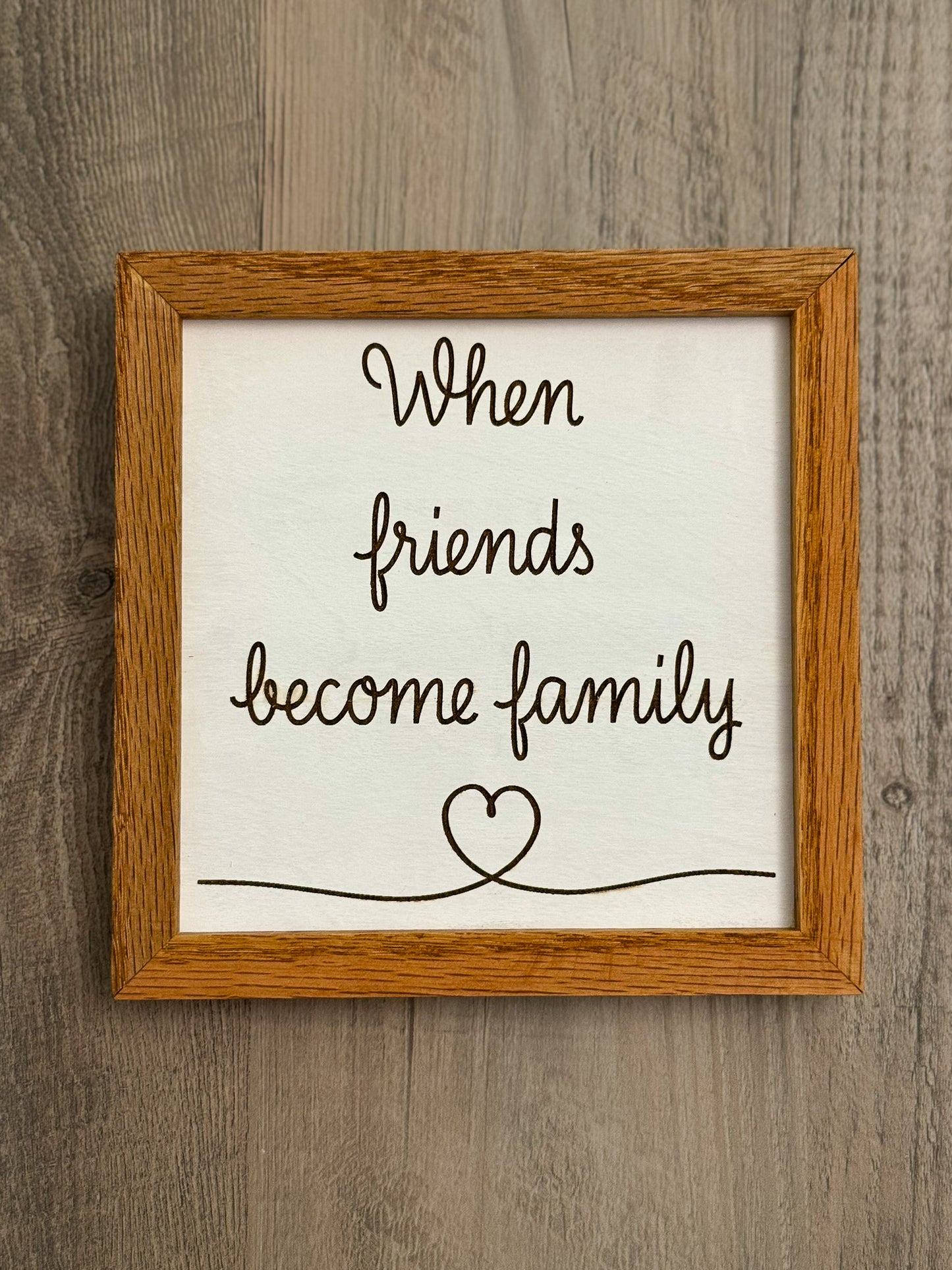 Square wooden sign framed with oak wood features a white painted background and laser engraved message "When friends become family" alongside a delicate heart design.