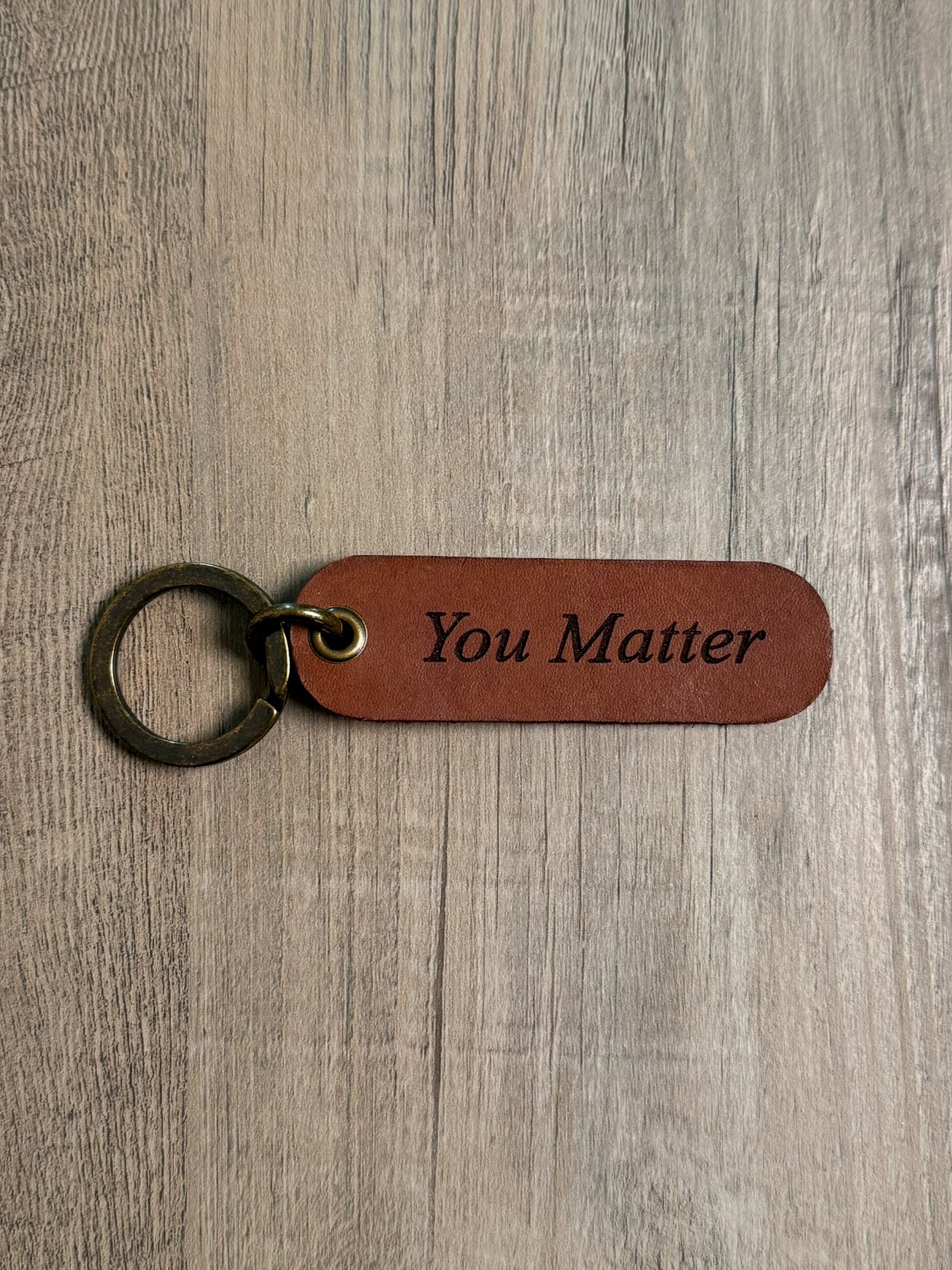 Brown rounded rectangle leather keychain with a brass key ring and jump ring. With the words "You Matter" laser-engraved on it. Approx. 1" X 3".