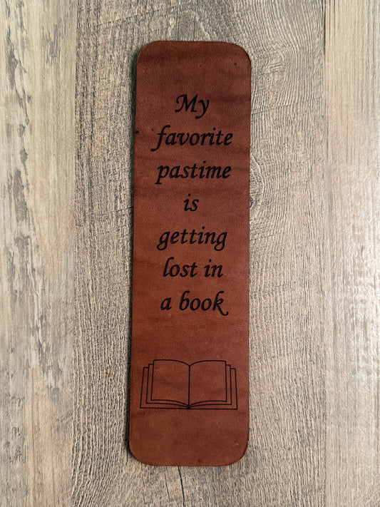 A brown, rectangular leather bookmark with rounded corners. Laser-engraved with the quote "My favorite pastime is getting lost in a book. " A laser-engraved outline of an open book is below the quote text. 