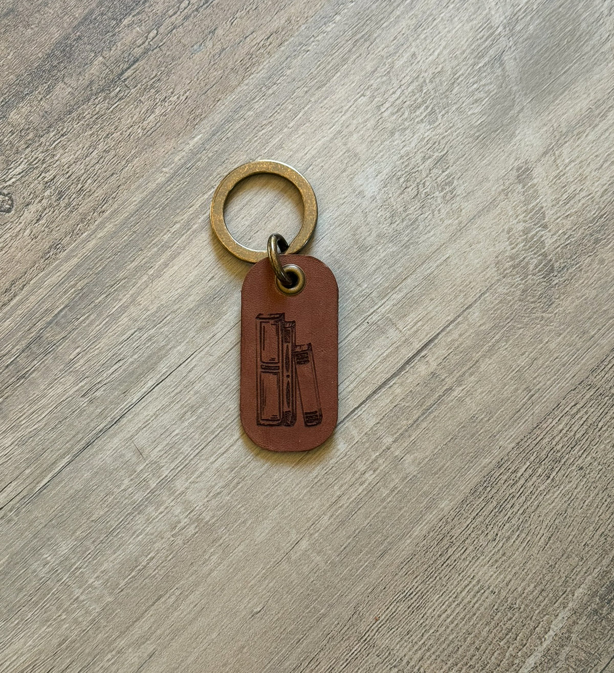 Brown rounded rectangle leather keychain with a brass key ring and jump ring. With books laser-engraved on it. Approx. 1" X 2". 