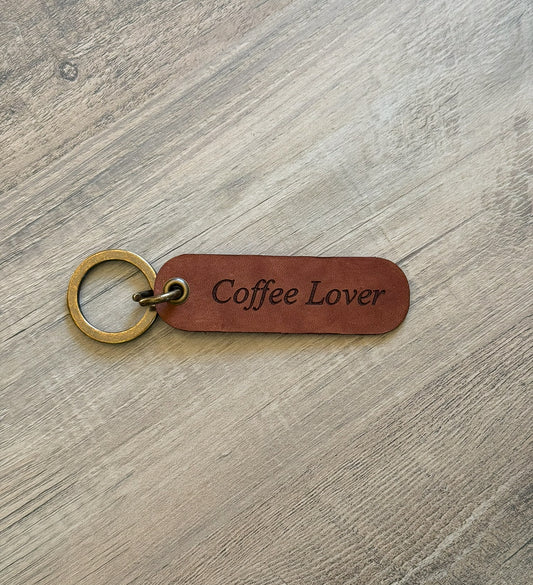 Brown rounded rectangle leather keychain with a brass key ring and jump ring. With the words "Coffee Lover" laser-engraved on it. Approx. 1" X 3". 