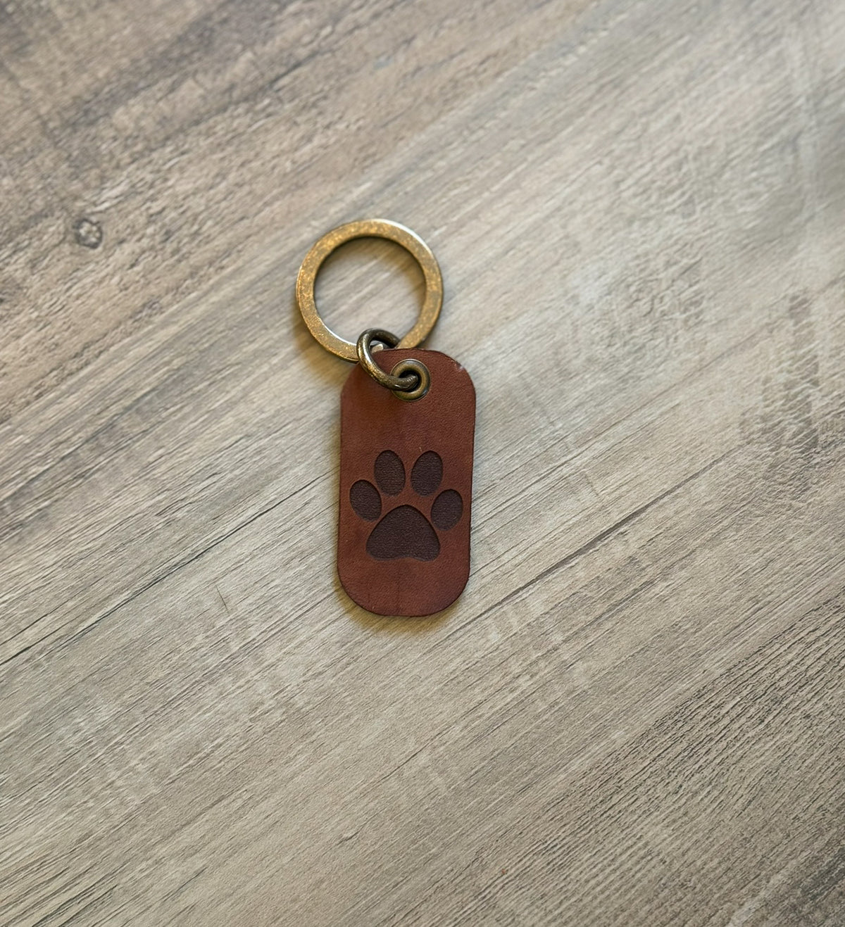 Brown rounded rectangle leather keychain with a brass key ring and jump ring. With a paw print laser-engraved on it. Approx. 1" X 2". 