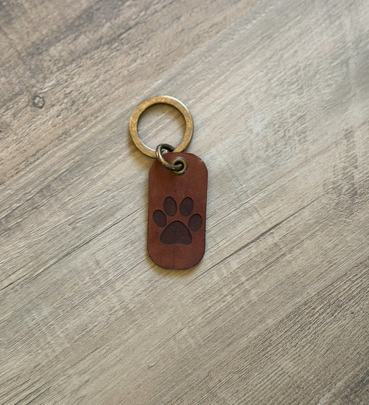 Brown rounded rectangle leather keychain with a brass key ring and jump ring. With a paw print laser-engraved on it. Approx. 1" X 2". 