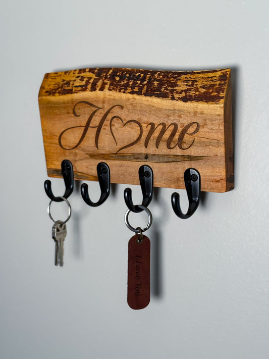 An ambrosia maple wood wall-mounted key holder with four black hooks, engraved with the word "Home" in cursive with the "o" being a heart.