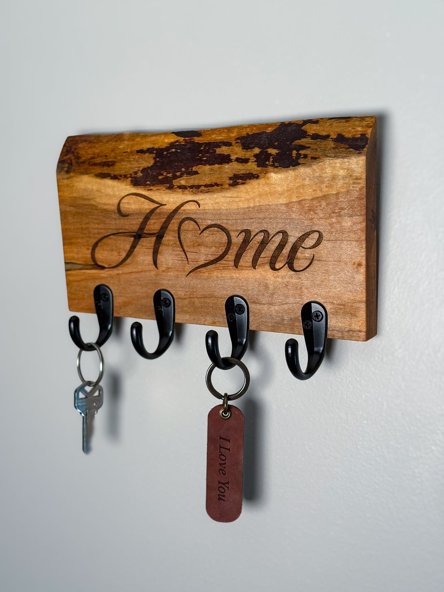 An ambrosia maple wood wall-mounted key holder with four black hooks, engraved with the word "Home" in cursive with the "o" being a heart.