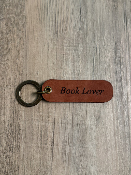 Brown rounded rectangle leather keychain with a brass key ring and jump ring. With the words "Book Lover" laser-engraved on it. Approx. 1" X 3". 