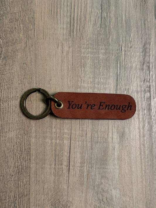 Brown rounded rectangle leather keychain with a brass key ring and jump ring. With the words "You're Enough" laser-engraved on it. Approx. 1" X 3". 