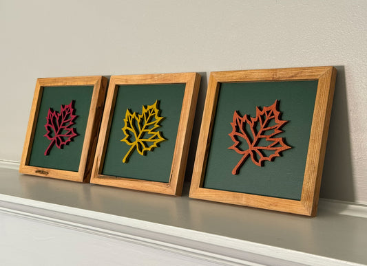 Three framed wooden leaf wall art pieces displayed side-by-side. Each square frame contains a laser-cut wooden maple leaf layered over a dark green background, with the frames in natural wood and the leaves in red, yellow, and orange tones.