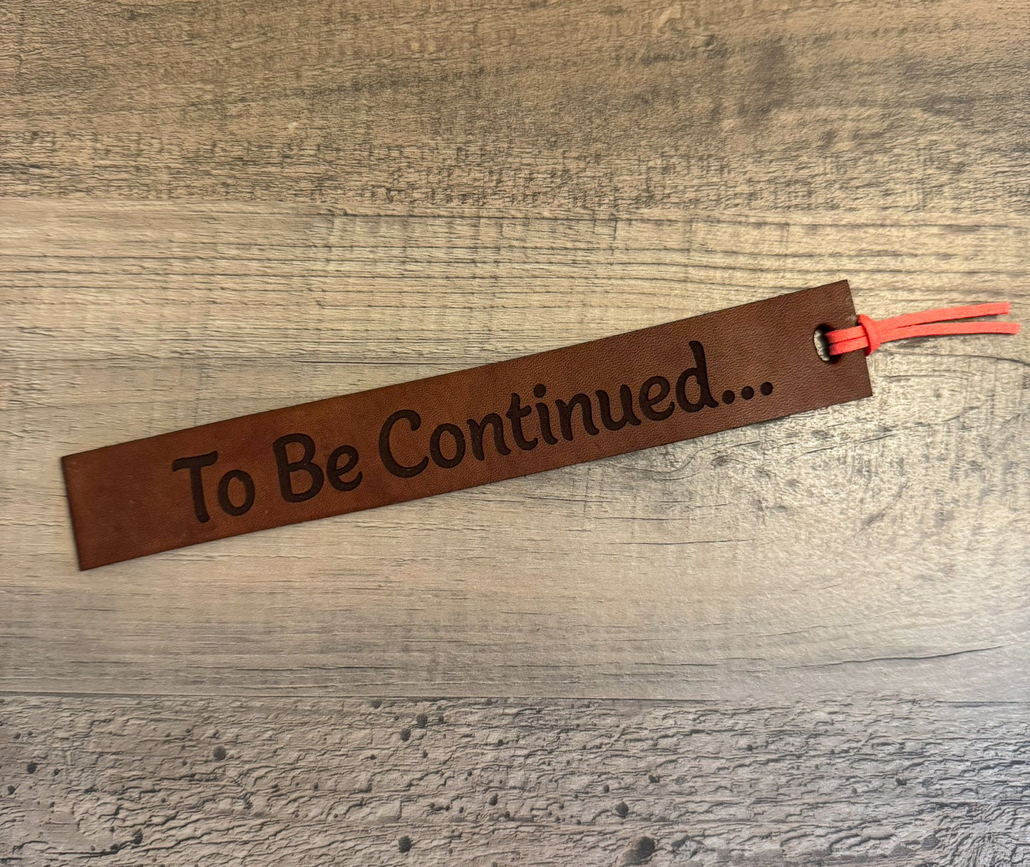 A brown, rectangular leather bookmark with a coral tassel laser-engraved with the quote "To Be Continued...".