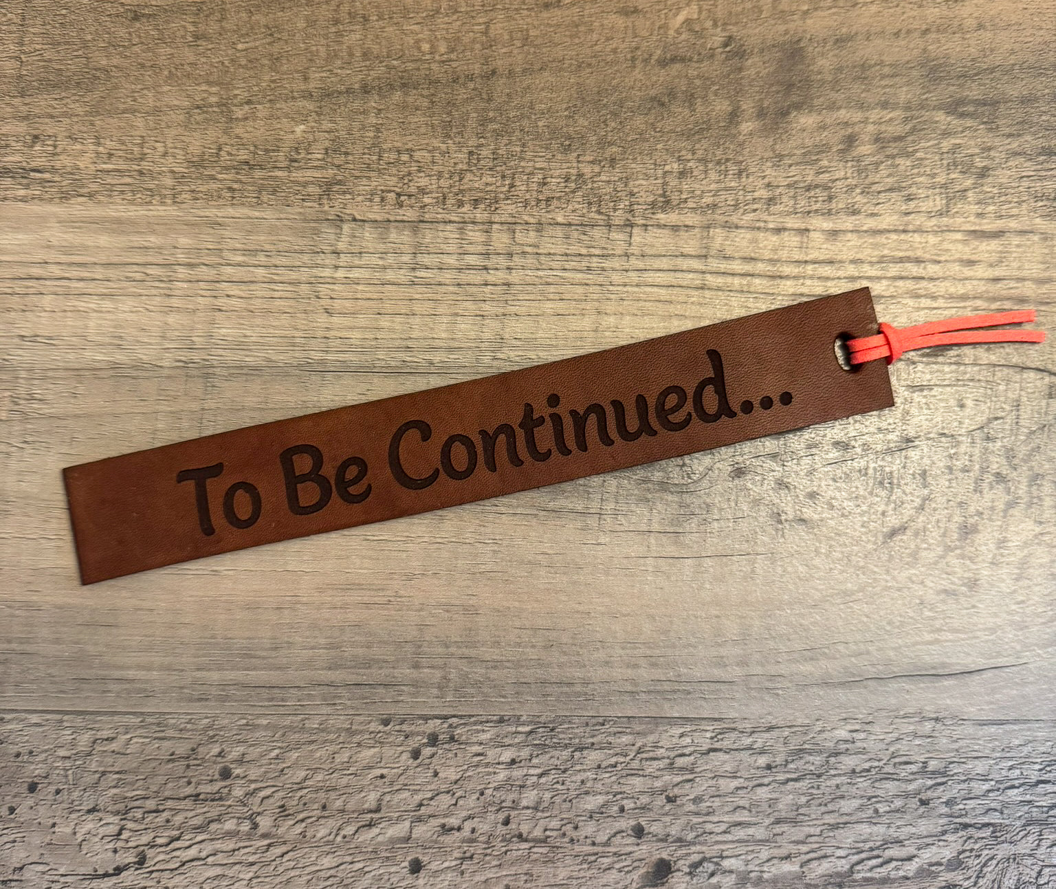 A brown, rectangular leather bookmark with a coral tassel laser-engraved with the quote "To Be Continued...".