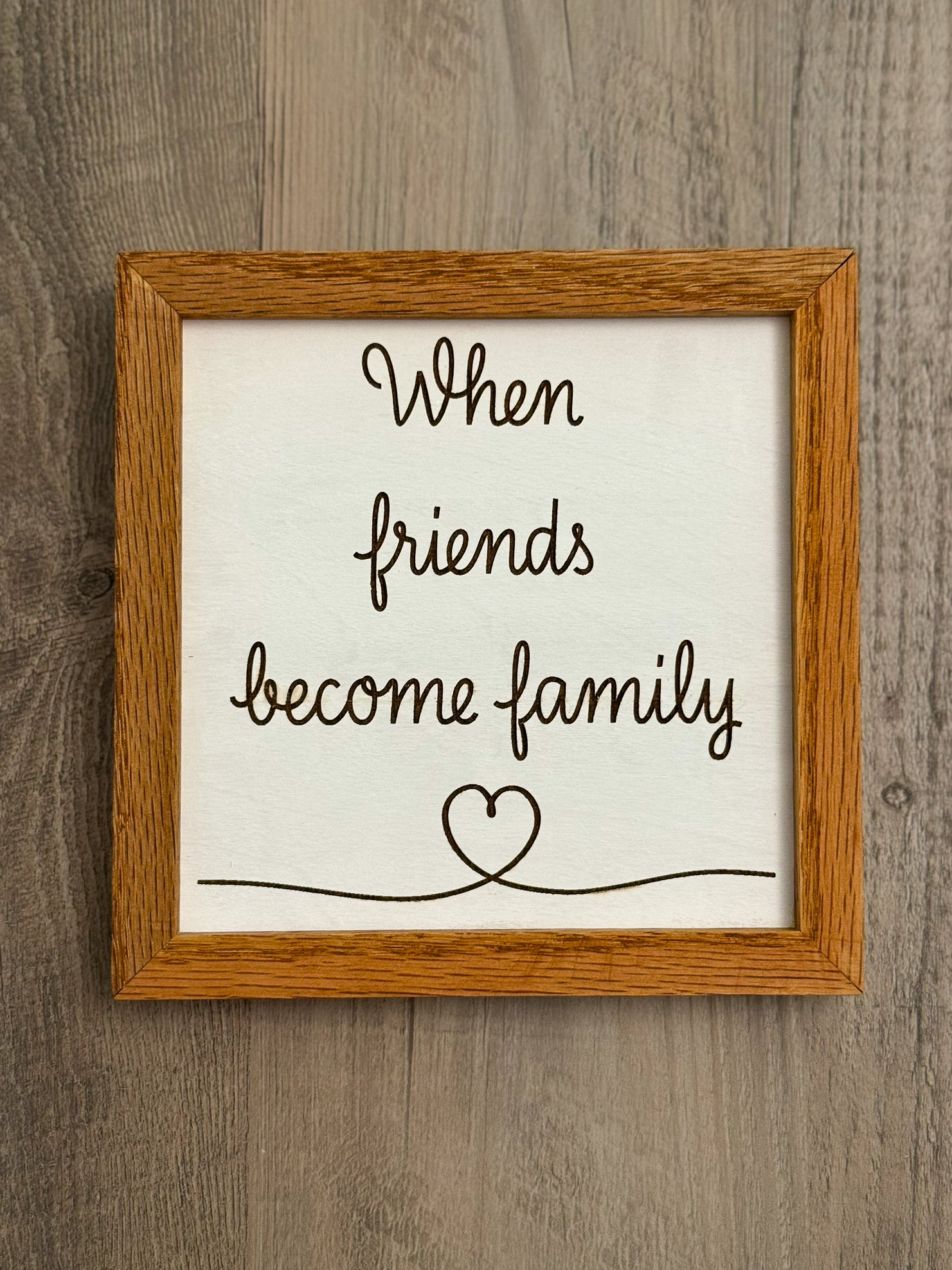 Square wooden sign framed with oak wood features a white painted background and laser engraved message "When friends become family" alongside a delicate heart design.