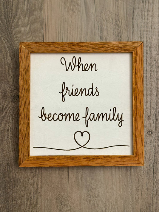 Square wooden sign framed with oak wood features a white painted background and laser engraved message "When friends become family" alongside a delicate heart design.