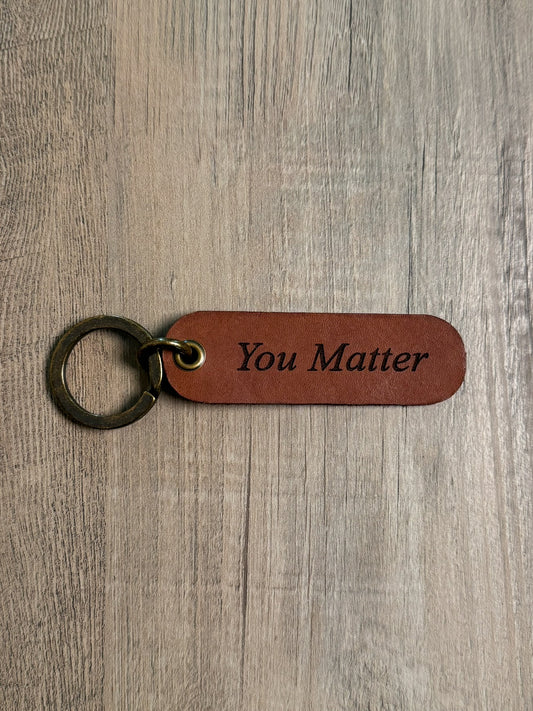 Brown rounded rectangle leather keychain with a brass key ring and jump ring. With the words "You Matter" laser-engraved on it. Approx. 1" X 3". 
